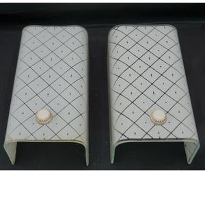 May include: A pair of rectangular, curved glass wall sconce shades featuring a white frosted finish with a black diamond grid pattern and small dots. Each shade includes a single, circular, off-white textured knob centered near the bottom.