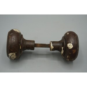 May include: An antique door knob set with a dark brown finish and white paint chipping. The set includes two round knobs connected by a metal rod. The knobs have a classic, vintage design.