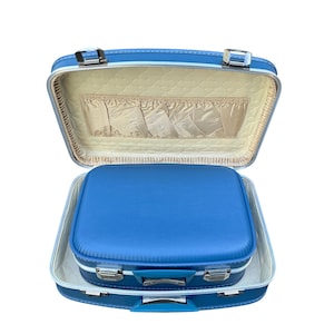 May include: A bright blue hard-shell suitcase with silver metal latches and hinges. The interior is lined with a cream-coloured quilted fabric featuring a gathered pocket. A smaller matching blue suitcase is nested inside the open main case.