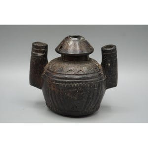 May include: A dark brown, rounded vessel with a central spout and two side handles. The surface features carved geometric patterns, including triangular motifs around the neck and vertical lines on the body, with visible signs of wear and texture.