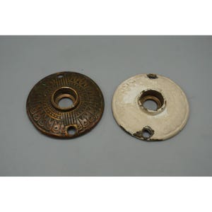 May include: Two antique door hardware rosettes. One is bronze-colored with an ornate, embossed design. The other is white with chipped paint. Both have a central hole and screw holes.