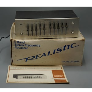 May include: A vintage Realistic 5-band stereo frequency equalizer. The silver-faced unit sits atop its original box, which also displays the Realistic logo and product information. An owner's manual is included. The unit has multiple sliders and knobs.
