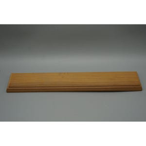 May include: A rectangular, light brown wooden shelf with a grooved surface. The shelf has a raised edge along the bottom and is set against a gray background. The wood grain is visible.