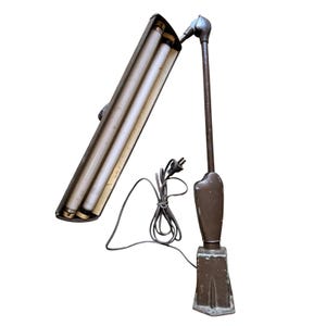 May include: An antique brown metal desk lamp with two fluorescent light tubes. The lamp has an adjustable arm and a rectangular base. The power cord is visible. The lamp is designed for task lighting.