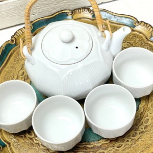 May include: A white ceramic teapot with a woven handle and four matching cups on a gold and green tray.
