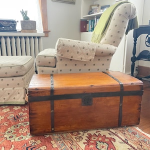 May include: A vintage wooden trunk with metal bands and a lock sits on a patterned rug. The trunk is rectangular and has a warm, brown finish. A patterned armchair and ottoman are in the background, along with a vintage typewriter and a window.