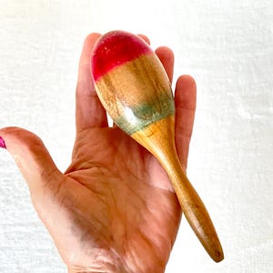 Pode incluir: Uma maraca de madeira com a parte superior vermelha e um acabamento em madeira natural. A maraca tem uma cabeça arredondada e um cabo longo e fino. A parte superior da maraca é pintada de vermelho e o resto do corpo é de cor de madeira natural.