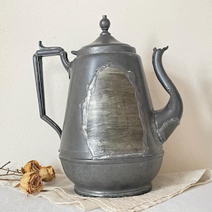 May include: Antique pewter coffee pot with a curved spout, a tall handle, and a lid with a small knob. The pot has a large, rectangular patch of lighter metal on the front. The pot is sitting on a white cloth with dried roses.