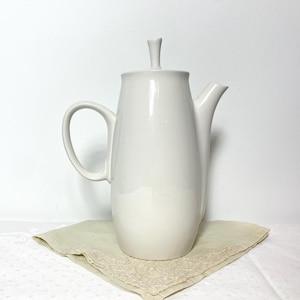 May include: A white ceramic coffee pot with a lid and a curved handle. The pot is placed on a beige cloth with a lace trim. The background is a plain white wall.