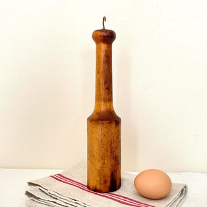 Puede incluir: Un mortero de madera vintage con un gancho de metal. El mortero es de color marrón claro y está sobre una toalla de lino con rayas rojas. Un huevo descansa junto al mortero. El mortero mide unos 25 cm de alto.