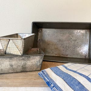 Puede incluir: Tres moldes para pan de metal vintage, uno apilado encima del otro, con un paño de cocina a rayas blancas y azules en el fondo.