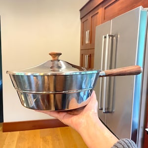 May include: A stainless steel saucepan with a polished finish, featuring a matching lid with a small wooden knob and a long, horizontal wooden handle attached to the side. The pan has a wide, circular shape with a slightly tapered base.