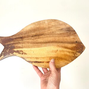 Puede incluir: Una tabla de cortar de madera con forma de pez y acabado natural de vetas. La tabla tiene una aleta caudal y un pequeño agujero cerca de la parte superior. La madera presenta tonos marrones y amarillos, con zonas más oscuras en los bordes.