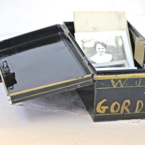 May include: A black metal box with a gold trim and the name "W.J. GORDON" written in gold lettering. The box is open and contains several photographs.