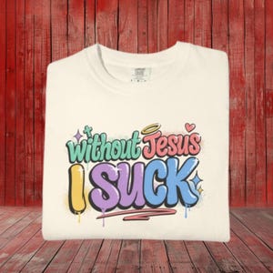 A cream-colored t-shirt is folded on a red wooden surface, displaying the colorful graffiti-style text "Without Jesus I Suck" on the front.
