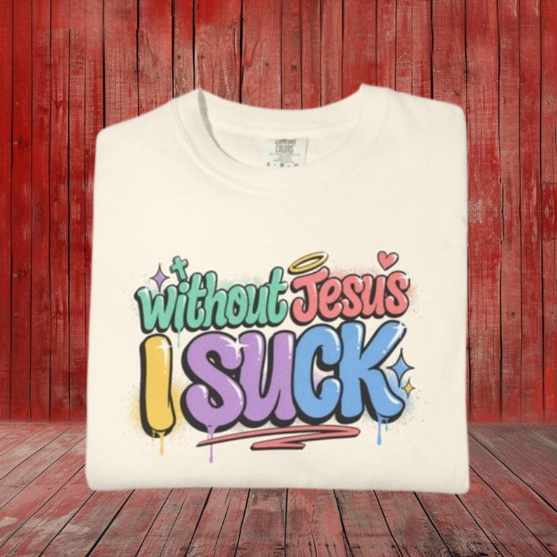 A cream-colored t-shirt is folded on a red wooden surface, displaying the colorful graffiti-style text "Without Jesus I Suck" on the front.