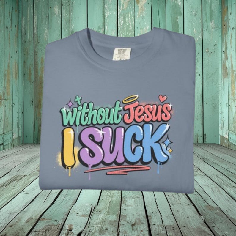 A light blue t-shirt is folded on a teal wooden surface, displaying the colorful graffiti-style text "Without Jesus I Suck" on the front.