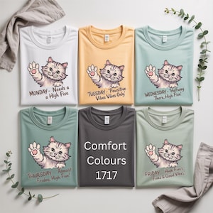 May include: A collection of six t-shirts in various colors, each featuring a cartoon cat waving. The shirts have text for each day of the week, including "Monday - Needs a High Five" and "Comfort Colours 1717".