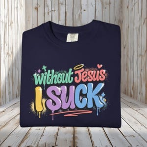 A navy blue t-shirt is folded on a light wooden surface, displaying the colorful graffiti-style text "Without Jesus I Suck" on the front.