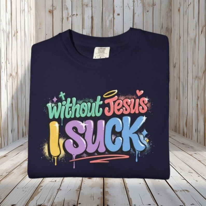 A navy blue t-shirt is folded on a light wooden surface, displaying the colorful graffiti-style text "Without Jesus I Suck" on the front.