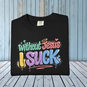 A black t-shirt is folded on a light wooden surface, displaying the colorful graffiti-style text "Without Jesus I Suck" on the front.