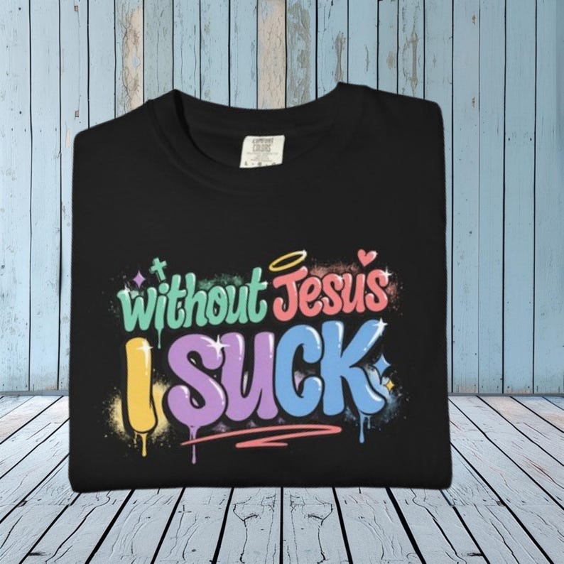 A black t-shirt is folded on a light wooden surface, displaying the colorful graffiti-style text "Without Jesus I Suck" on the front.