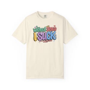 A cream-colored t-shirt with the colorful graffiti-style text "Without Jesus I Suck" is laid flat against a white background.