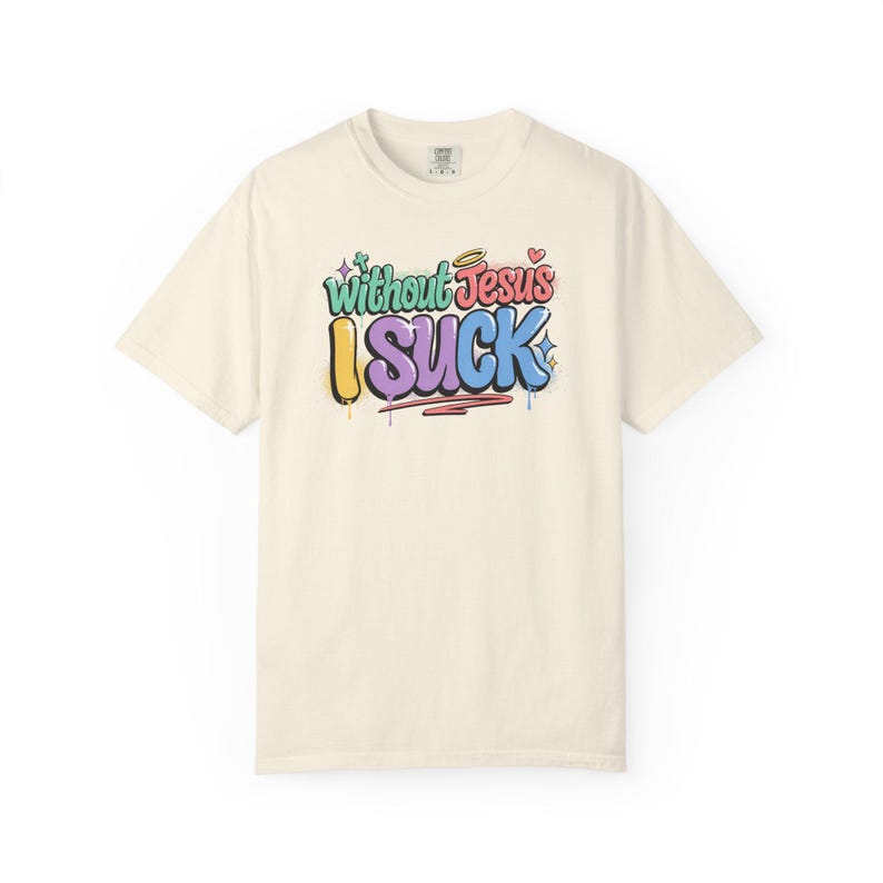 A cream-colored t-shirt with the colorful graffiti-style text "Without Jesus I Suck" is laid flat against a white background.