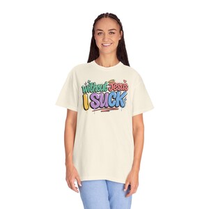 A woman with braided hair smiles while wearing an off-white t-shirt with "Without Jesus I Suck" in colorful, graffiti-style text.