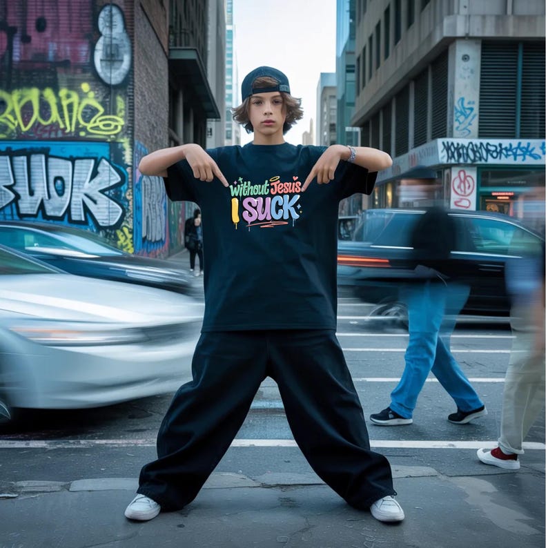 A young person in a black t-shirt with colorful graffiti-style text "Without Jesus I Suck" points to the text while standing on a city street.