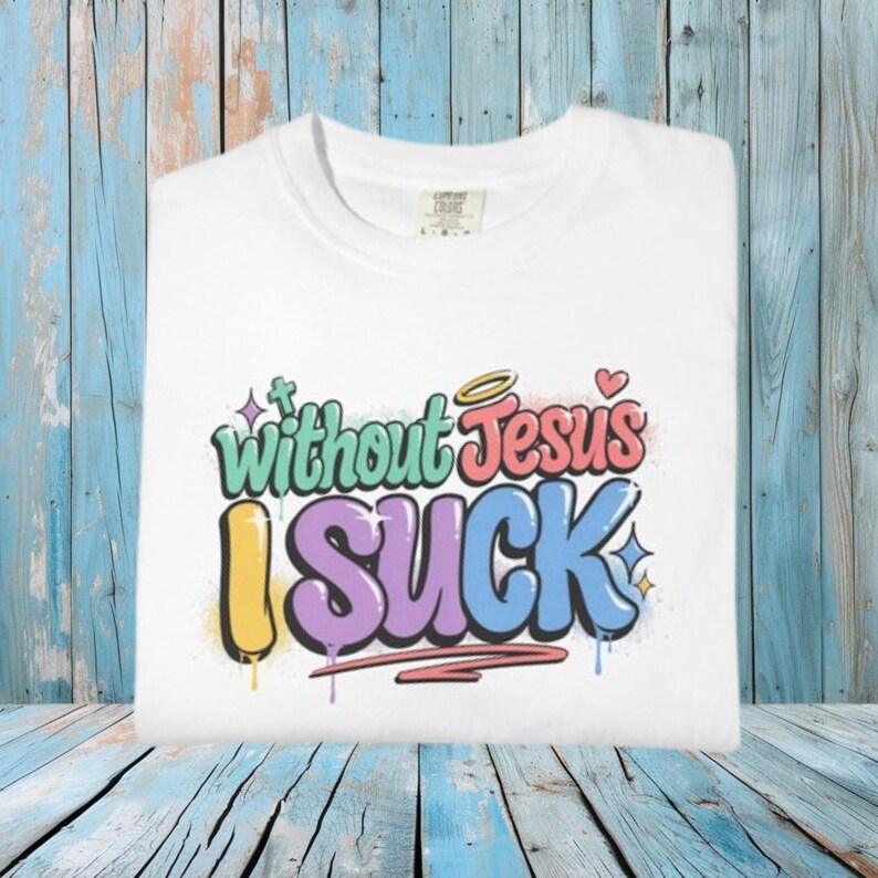 A white t-shirt is folded on a blue wooden surface, displaying the colorful graffiti-style text "Without Jesus I Suck" on the front.