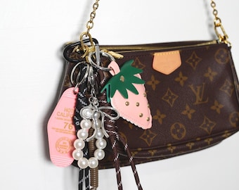 May include: A brown leather purse with a gold chain strap and a variety of charms attached, including a pink strawberry, a pearl necklace, and a pink motel key chain with the text "Hotel California 76".
