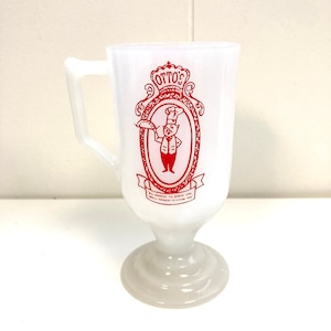 May include: White ceramic mug with a red logo that says "Otto's" and features a cartoon chef holding a pizza. The logo is surrounded by a decorative frame. The mug has a handle and a pedestal base.