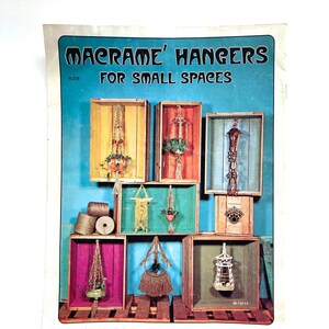 May include: A vintage craft book cover with the title "Macrame' Hangers For Small Spaces". The cover features a colorful collage of macrame plant hangers displayed in wooden crates against a blue background.