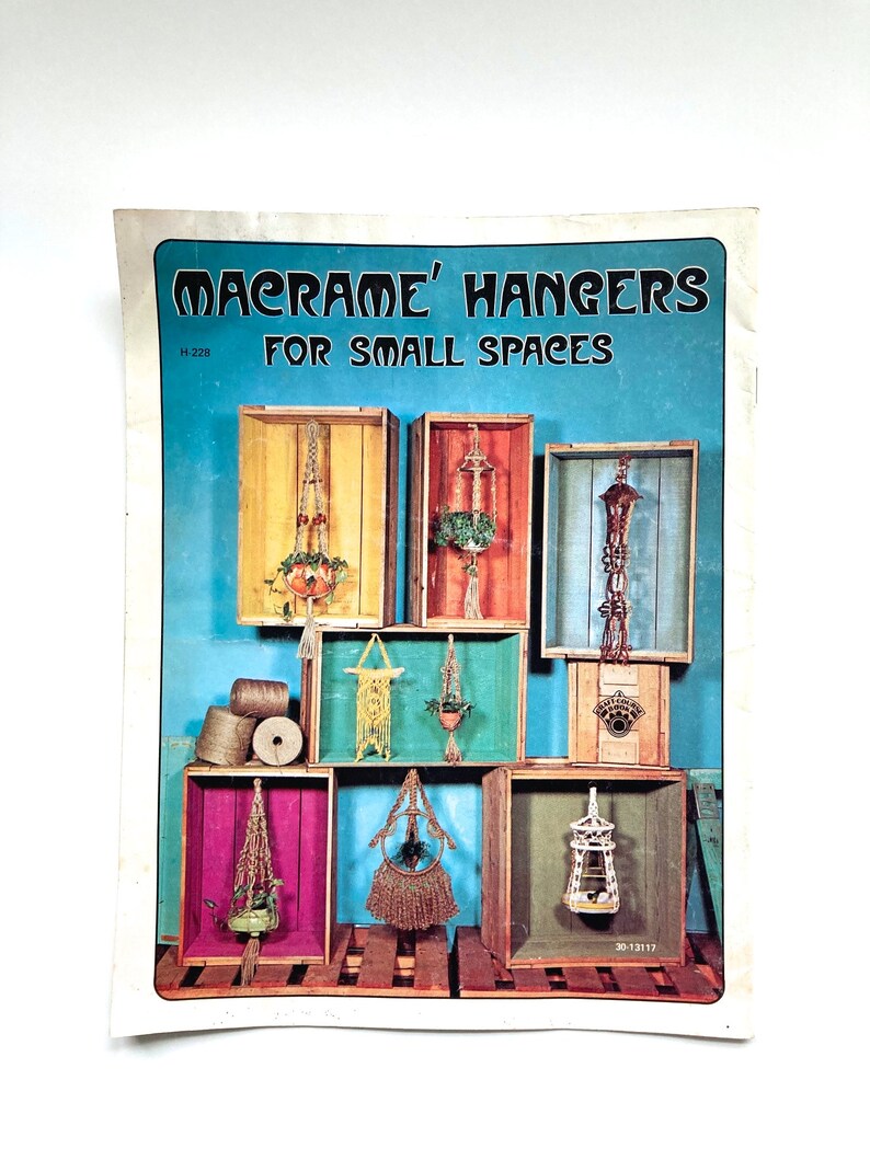 May include: A vintage craft book cover with the title "Macrame' Hangers For Small Spaces". The cover features a colorful collage of macrame plant hangers displayed in wooden crates against a blue background.