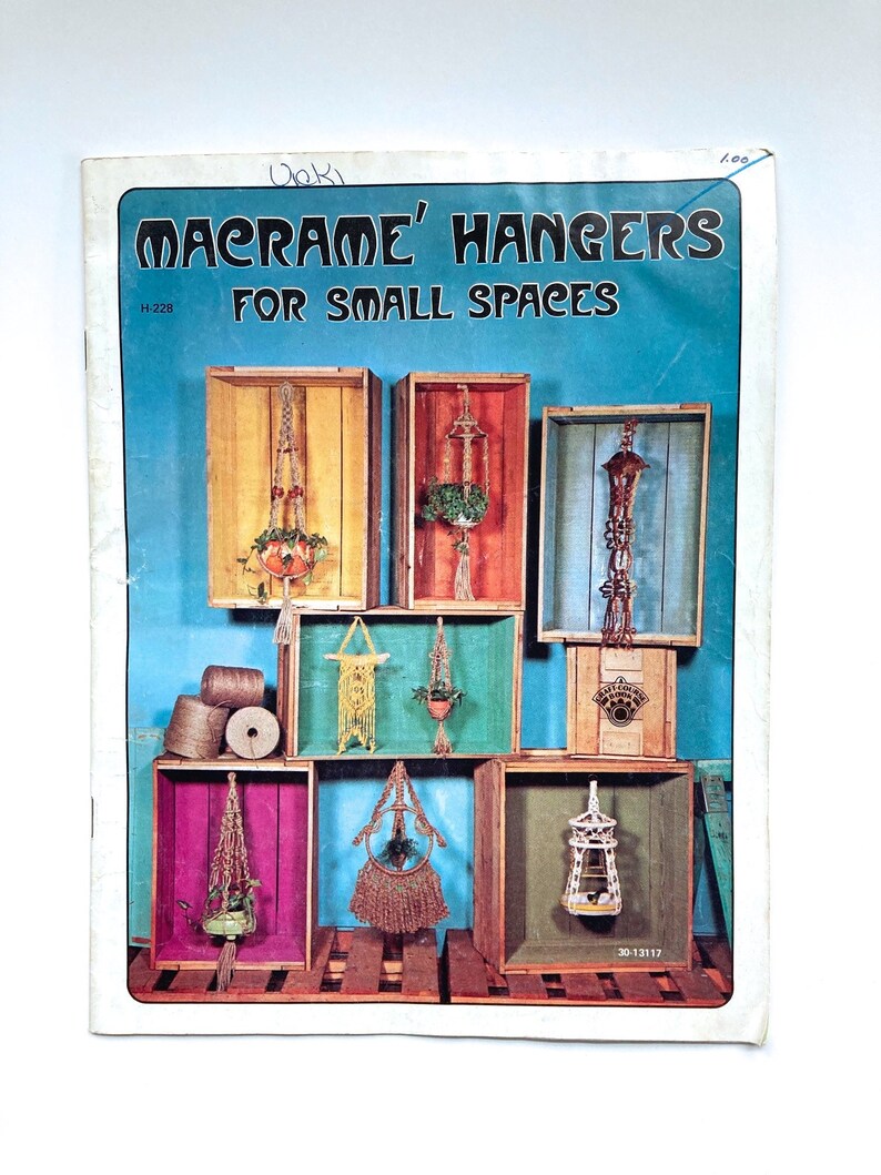 May include: A vintage book cover with the title "Macrame' Hangers For Small Spaces" and a photo of macrame plant hangers displayed in wooden crates.