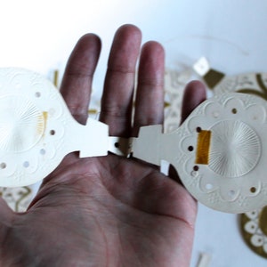 May include: Two white paper Christmas ornaments with a gold thread detail. The ornaments are open and have a delicate, embossed design.