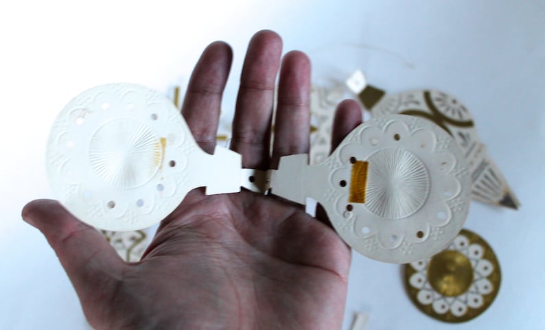 May include: Two white paper Christmas ornaments with a gold thread detail. The ornaments are open and have a delicate, embossed design.