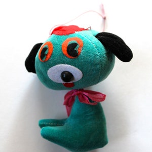 May include: A teal blue plush toy dog with black ears, orange eyes, and a red and white nose. The dog has a pink bow around its neck.