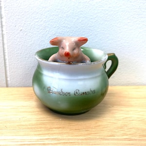 May include: A small, vintage ceramic cup with a green and white ombre design, featuring a handle and gold trim. A small, pink pig figurine is inside the cup. The text "Windsor Canada" is written on the cup.