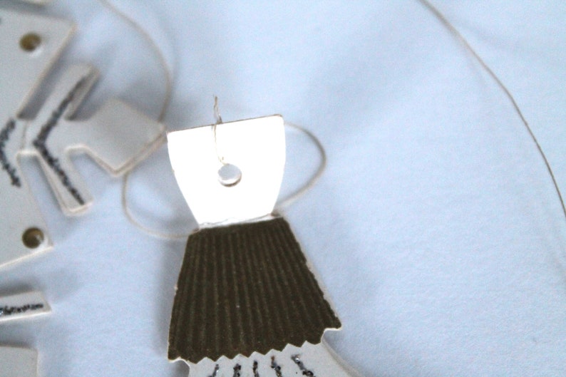 May include: A white paper doll with a brown paper skirt and a hole in the top for hanging. The doll is hanging from a string.