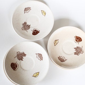 May include: Three white ceramic saucers with a brown and yellow leaf design. The saucers have a speckled texture.