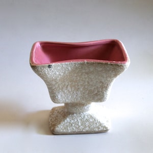 May include: A white ceramic planter with a pink glazed interior. The planter has a textured, rough surface and a square base.