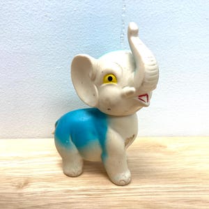 May include: A vintage toy elephant figurine. The elephant is primarily off-white with a blue back and yellow eyes. The trunk is raised, and the mouth is open with a red interior. The toy is made of a hard plastic material.