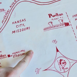 May include: A red and white map of Kansas City, Missouri, with a red star and the text "Puritan 20 the Grand" in the center. The map shows the locations of the Municipal Auditorium, Municipal Stadium, North Kansas City, Liberty Memorial, and the Missouri River. The map also shows the locations of two Puritan plants, one in Kansas City and one in Independence, Missouri. The map also shows the location of the Santa Fe Railroad.