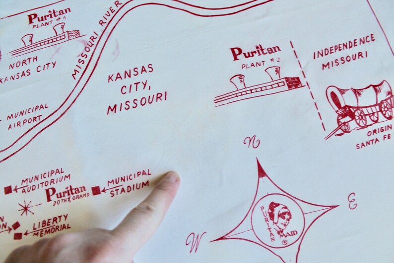 May include: A red and white map of Kansas City, Missouri, with a red star and the text "Puritan 20 the Grand" in the center. The map shows the locations of the Municipal Auditorium, Municipal Stadium, North Kansas City, Liberty Memorial, and the Missouri River. The map also shows the locations of two Puritan plants, one in Kansas City and one in Independence, Missouri. The map also shows the location of the Santa Fe Railroad.