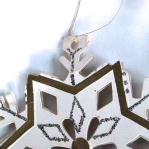 May include: White paper snowflake ornament with gold trim and silver glitter accents. The snowflake has a loop for hanging.