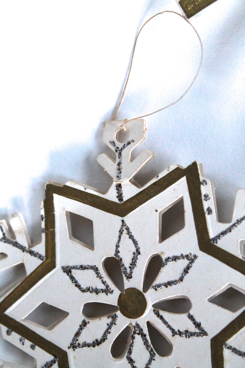 May include: White paper snowflake ornament with gold trim and silver glitter accents. The snowflake has a loop for hanging.