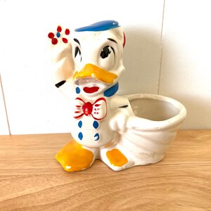 May include: A ceramic Donald Duck planter figurine. The planter is white with yellow, red, and blue accents. Donald Duck is holding a flower and wearing a bow tie. The planter has a small opening for plants.
