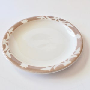 May include: A white ceramic plate with a brown border featuring a repeating white floral design. The plate is round and has a glossy finish, suitable for serving food. This plate is ideal for everyday use or special occasions.